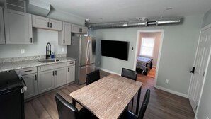 A modern kitchen with grey cabinets, a stainless steel refrigerator, and a dining area with a wooden table and chairs. - Almira Properties Unit 3 (Providence)