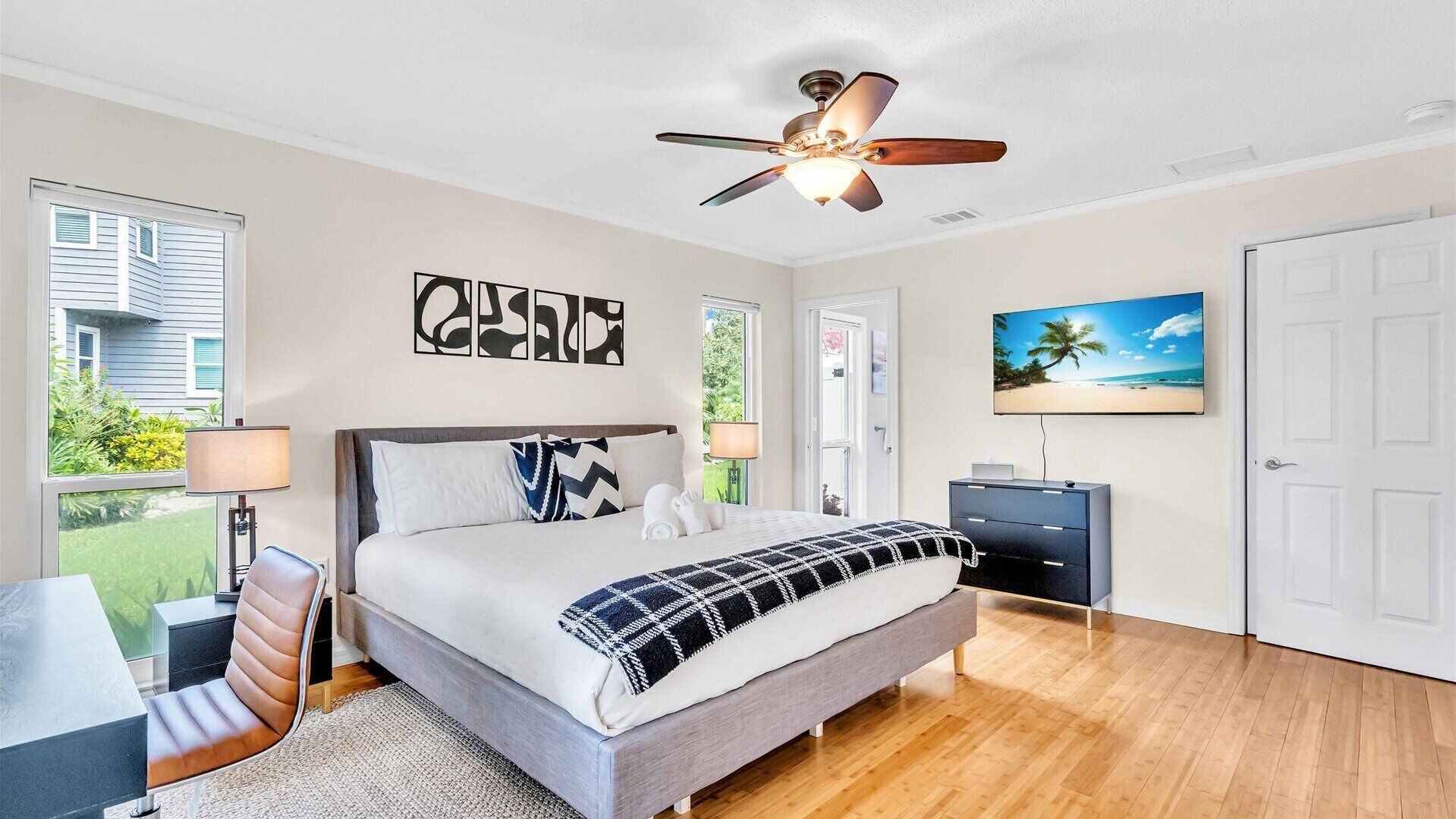 A bedroom with a bed, bedside tables, a desk, a chair, a ceiling fan, a TV, and a door.