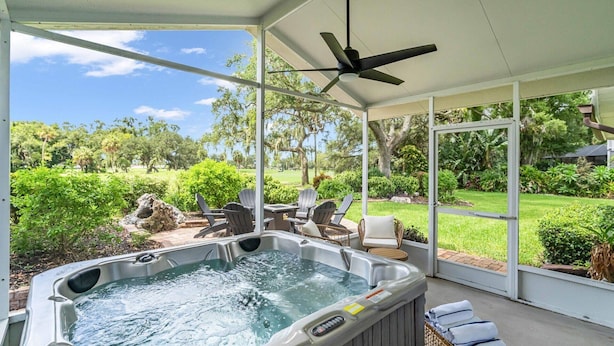 Un área al aire libre con jacuzzi cubierto, sillas y una mesa, rodeada de vegetación exuberante y vistas a un campo de golf.