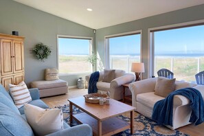 A spacious living room with a view of the beach, featuring a wooden cabinet, a blue sofa, a beige armchair, and a coffee table. - Beautiful Ocean Front Home! Sweeping Ocean Views! (Waldport)