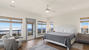 A bedroom with a large bed, a chair, a nightstand, and a view of the ocean through large windows.
