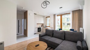 Una sala moderna con un sofá gris, una mesa de centro de madera y un control remoto encima. Se aprecia una cocina al fondo con armarios blancos y una ventana con cortinas.