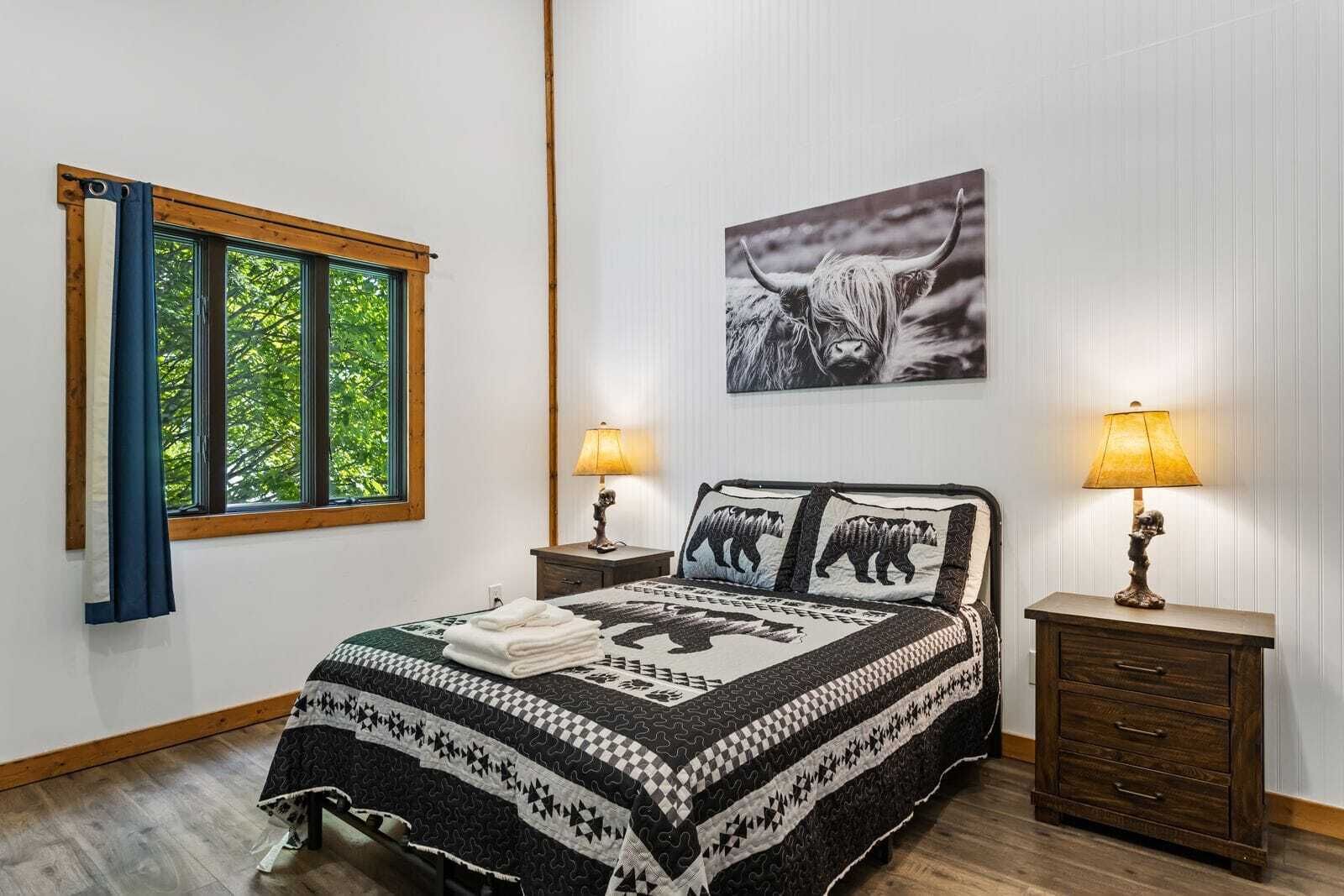 Une chambre à coucher comprenant un lit, deux tables de chevet, une fenêtre avec des rideaux, une lampe fixée au mur et un tableau encadré.