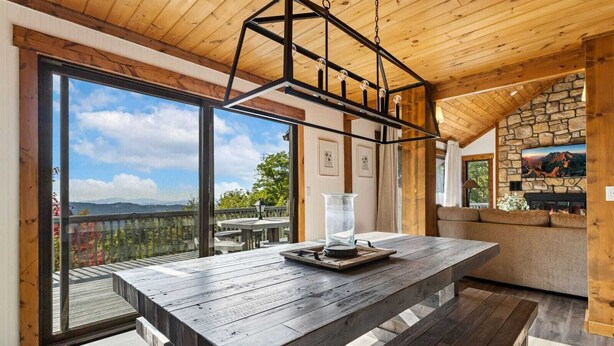A spacious room with a large wooden table, a stone fireplace, and a balcony with a scenic mountain view.