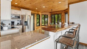 A modern kitchen with wooden ceiling beams, a granite countertop, stainless steel appliances, and a bar area with metal chairs.