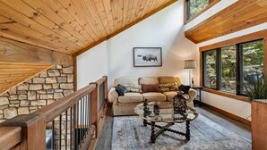 A cozy living room with a stone staircase, wooden ceiling, and large windows offering a view of the outdoors.