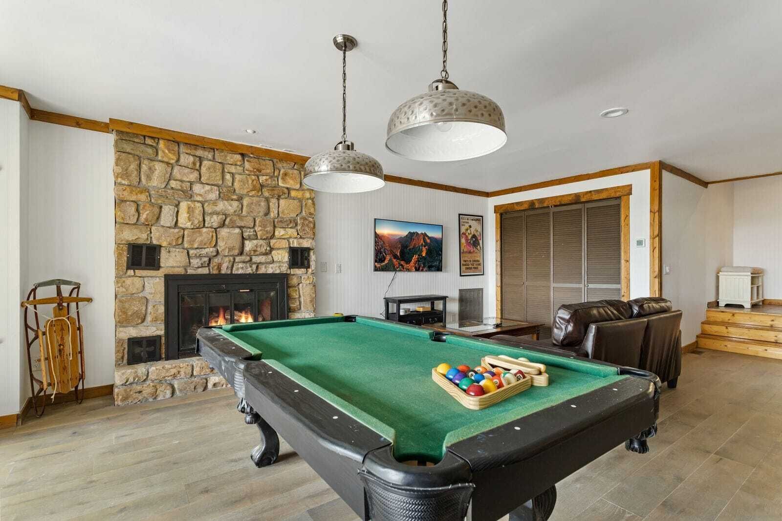 A pool table with a green felt surface, a stone fireplace, and a hanging light fixture.