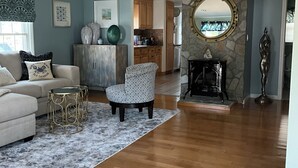A spacious living room with a sofa, armchair, and coffee table, a kitchen in the background, and a stone fireplace. - Charming year-round beach home in Indian Town, Old Saybrook (Old Saybrook)