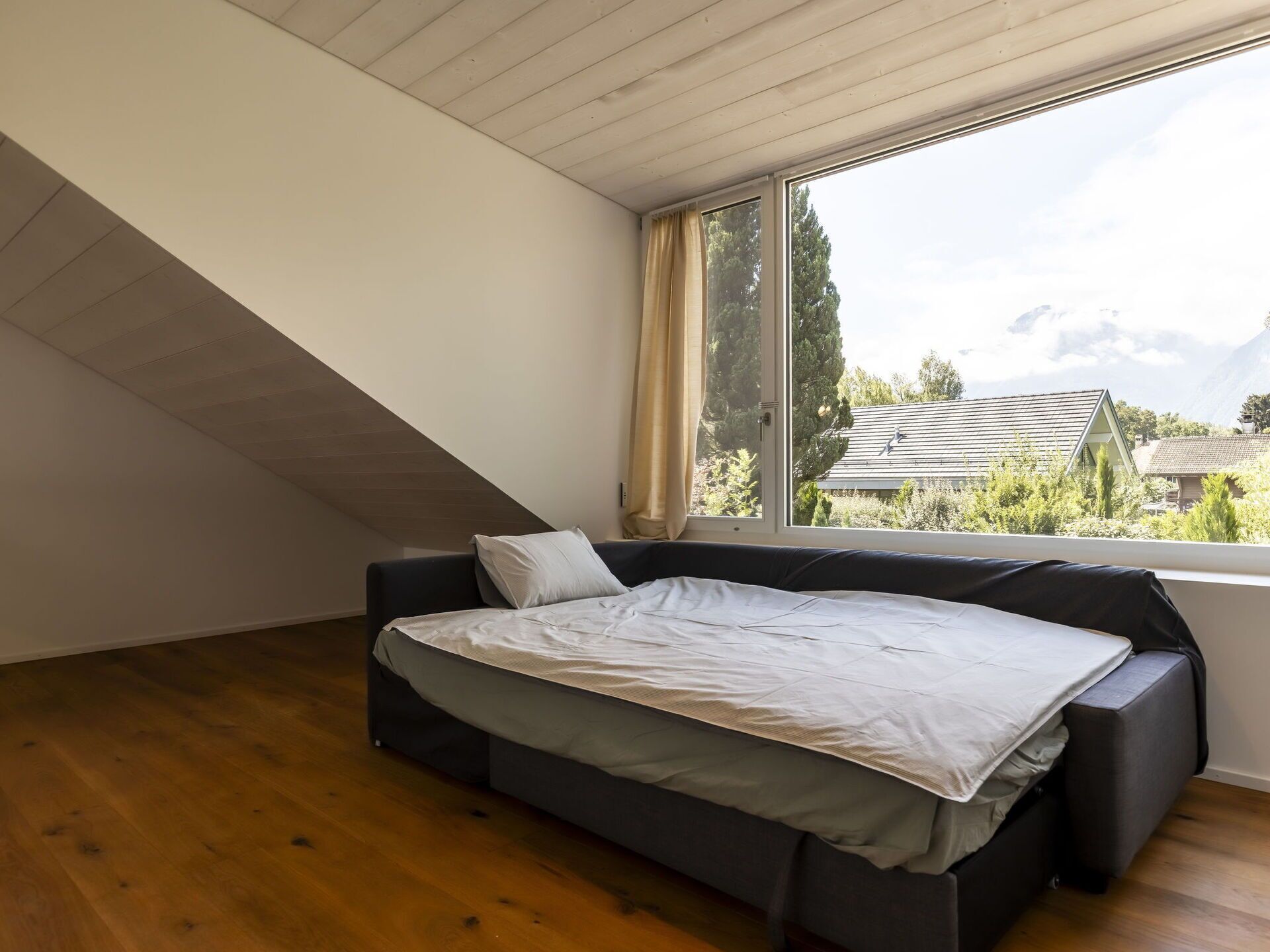 A bedroom with a bed, a window with a view of houses and trees, and a sloped ceiling.