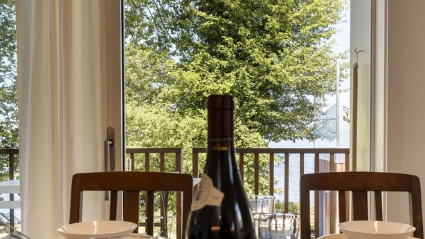 Ein Esstisch mit einer Weinflasche, Gläsern und Tellern, vor einer Kulisse aus einem Balkon mit Blick auf Bäume und ein Gewässer.