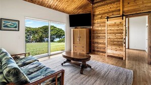 Ein geräumiges Wohnzimmer mit Holzboden, einem großen Schiebeglasfenster, das zu einer grünen Außenfläche führt, einem Holzschrank und einem an der Wand befestigten Flachbildfernseher.