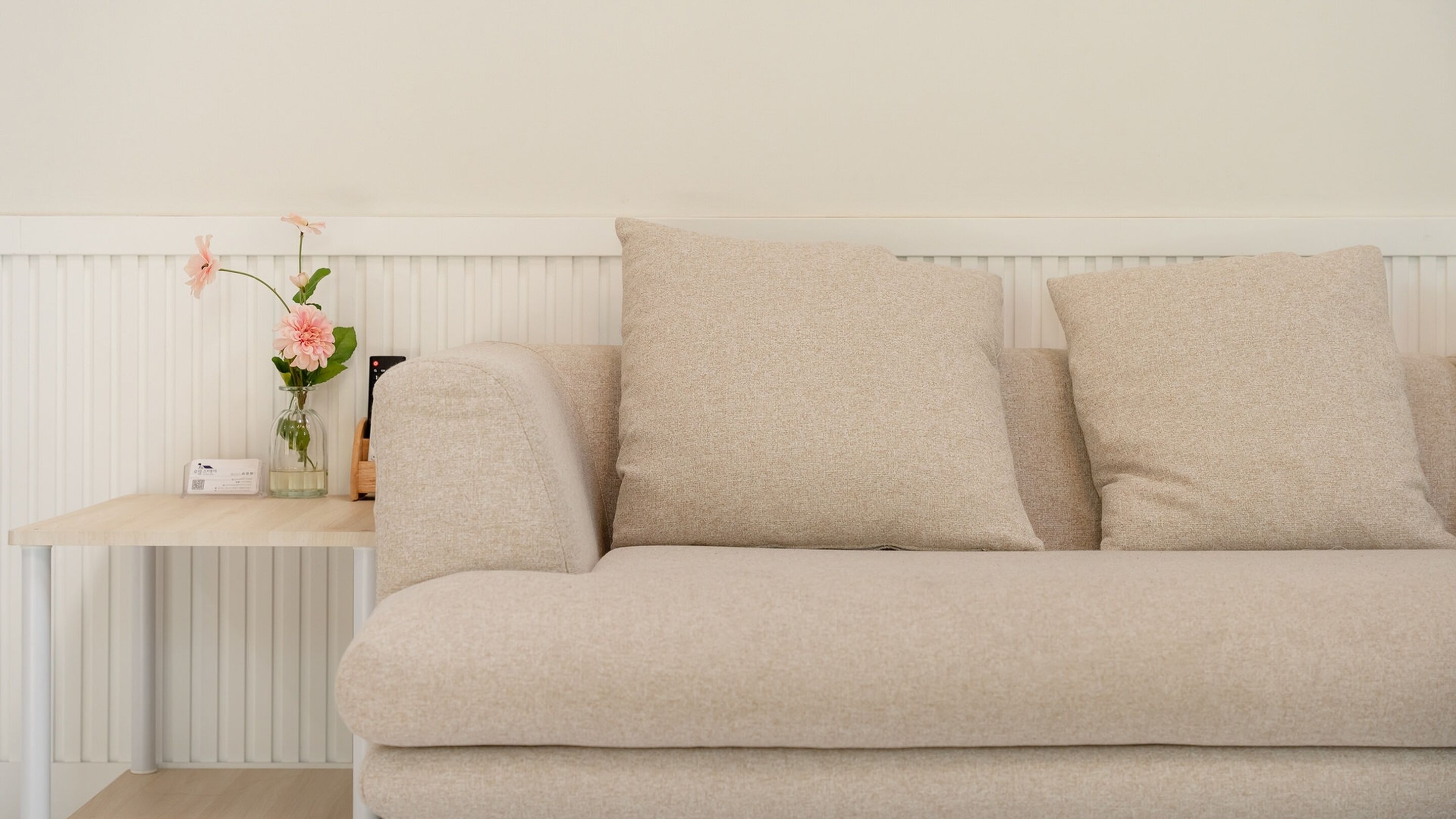 En beige soffa med två kuddar mot en vit vägg, ett litet bord med en vas med blommor och en fjärrkontroll.