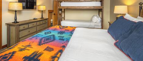 Una habitación de hotel con literas, un edredón colorido, un aparador de madera y dos lámparas.