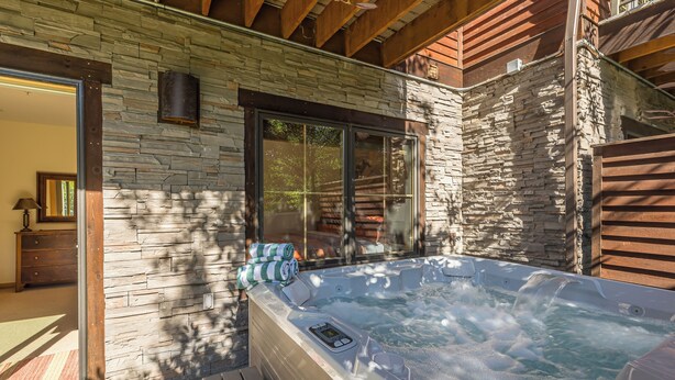 A hot tub with a stone exterior wall and wooden beams, a wooden deck, and a glass door leading to an interior room. - Private hot tub, multiple private decks, near Town Center - Spanish Peaks Perfection (Big Sky)