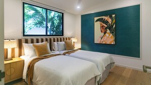 Quarto com uma cama grande, duas mesas de cabeceira, um ventilador de teto, uma janela com vista para palmeiras e um quadro na parede.
