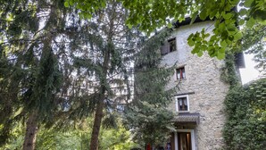 Un bâtiment en pierre avec une porte et des fenêtres en bois, entouré d’arbres et bordé d’un sentier menant à un banc.