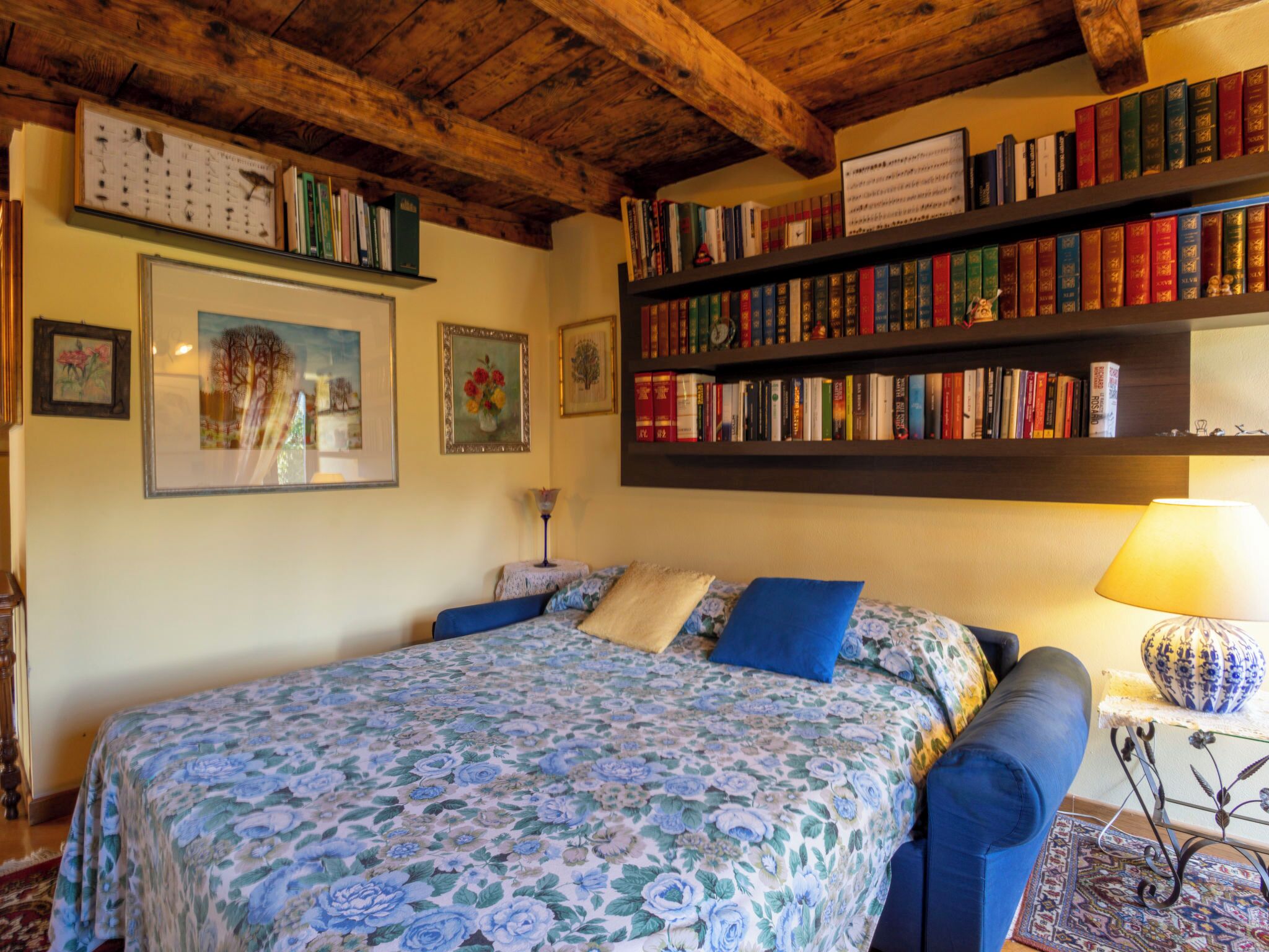 A bedroom with a floral bedspread, a bookshelf, and framed artwork on the wall.