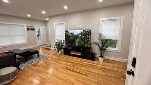 Een moderne woonkamer met een flatscreen televisie, houten vloeren, een zwarte tv-meubel en een potplant.