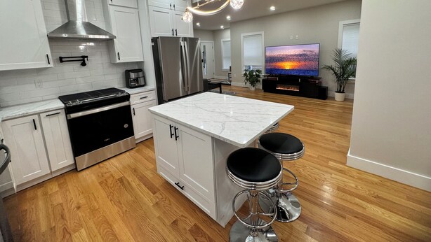 Een moderne keuken met witte kasten, roestvrijstalen apparatuur, een marmeren eiland en een grote televisie op de achtergrond.