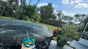 A person relaxing on a deck with a car covered by a tarp, a potted plant, and a clear sky.