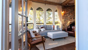 A cozy living room with a stone fireplace, a grey sectional sofa, and a wooden chair. - Marin Castle: Private Resort w Top of World Views, Hot Tub & Mini Golf, 30 Acres (Lagunitas)