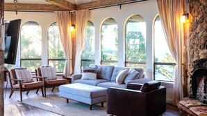 A cozy living room with a sectional sofa, armchairs, and a fireplace. Large windows with curtains provide natural light and views of the outdoors. - Marin Castle: Private Resort w Top of World Views, Hot Tub & Mini Golf, 30 Acres (Lagunitas)