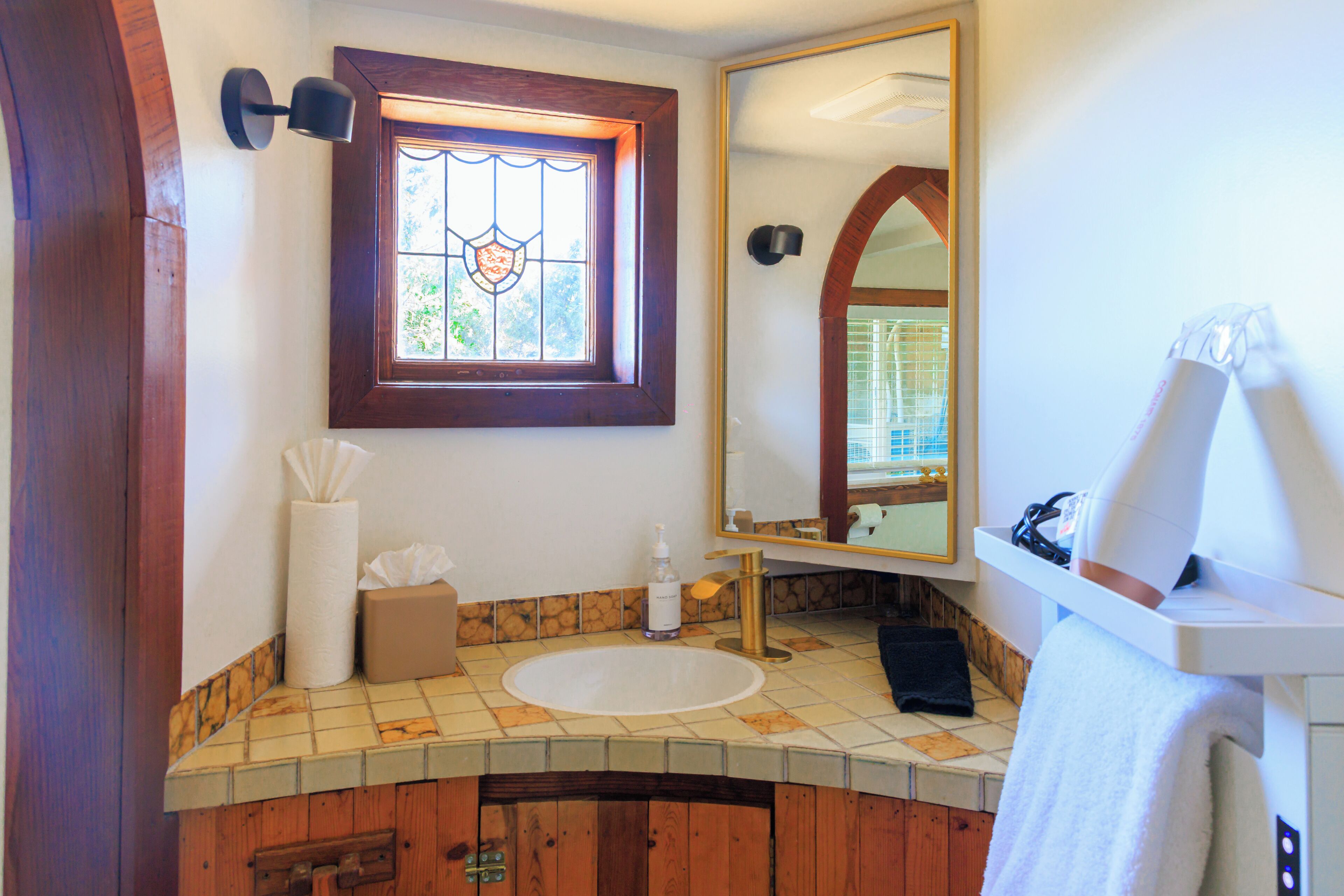 Un baño con tocador de madera, un espejo grande, una ventana con vidrieras y un lavamanos blanco.