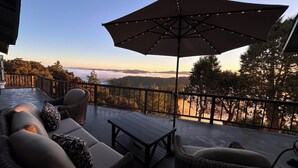 A balcony with outdoor furniture, a large umbrella, and a view of a forest and mountains. - Marin Castle: Private Resort w Top of World Views, Hot Tub & Mini Golf, 30 Acres (Lagunitas)