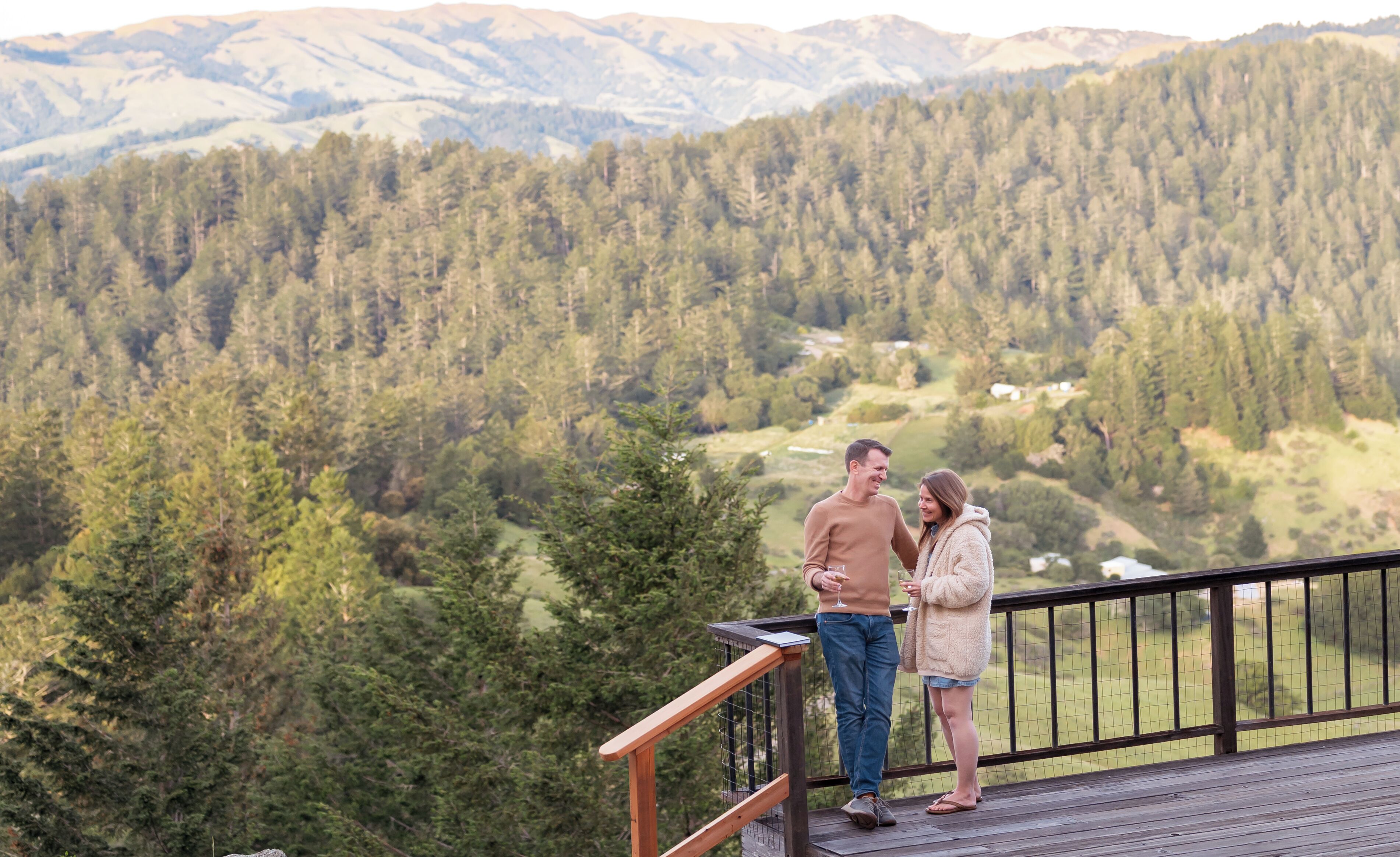 Una pareja en una terraza de madera con vistas a un valle boscoso y montañas al fondo.