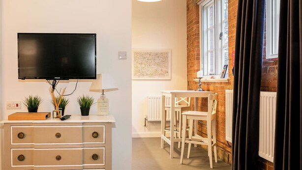 Una habitación con televisión, cómoda, pequeña mesa con sillas, radiador y ventana con cortinas.