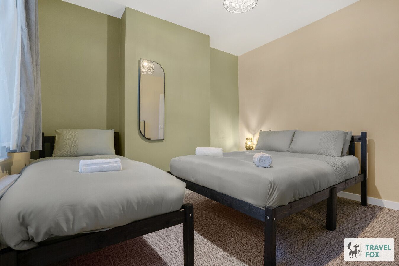 A hotel room with two beds, a mirror, a nightstand with a lamp, and a carpeted floor.
