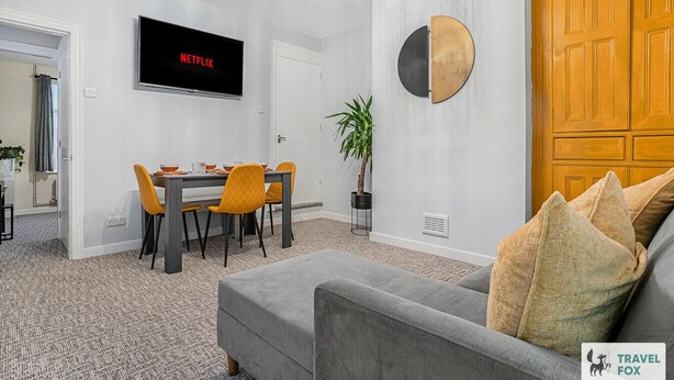 A modern living room with a grey sofa, a dining table with orange chairs, a TV displaying 'NETFLIX', and a wooden door.