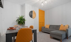 Ein modernes Wohnzimmer mit einem grauen Sofa, orangefarbenen Kissen und einem Essbereich mit Tisch für zwei Personen.