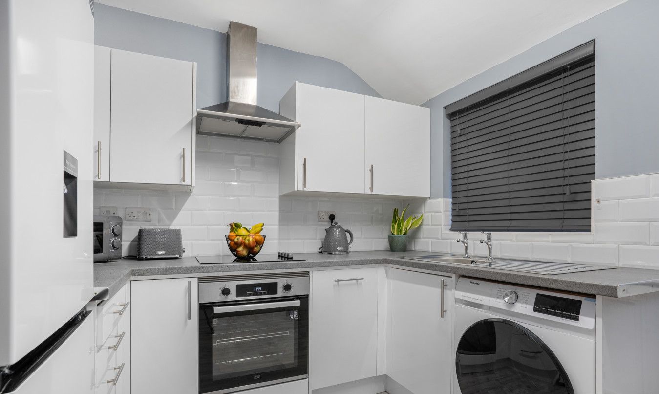 A modern kitchen with white cabinets, a built-in oven, a washing machine, and a window with blinds.