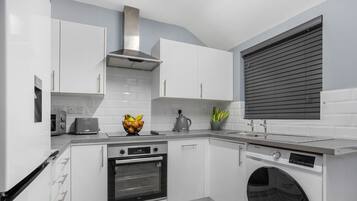 A modern kitchen with white cabinets, a built-in oven, a washing machine, and a window with blinds.