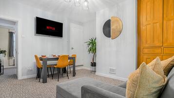 A modern living room with a grey sofa, a dining table with orange chairs, a TV displaying 'NETFLIX', and a wooden door.