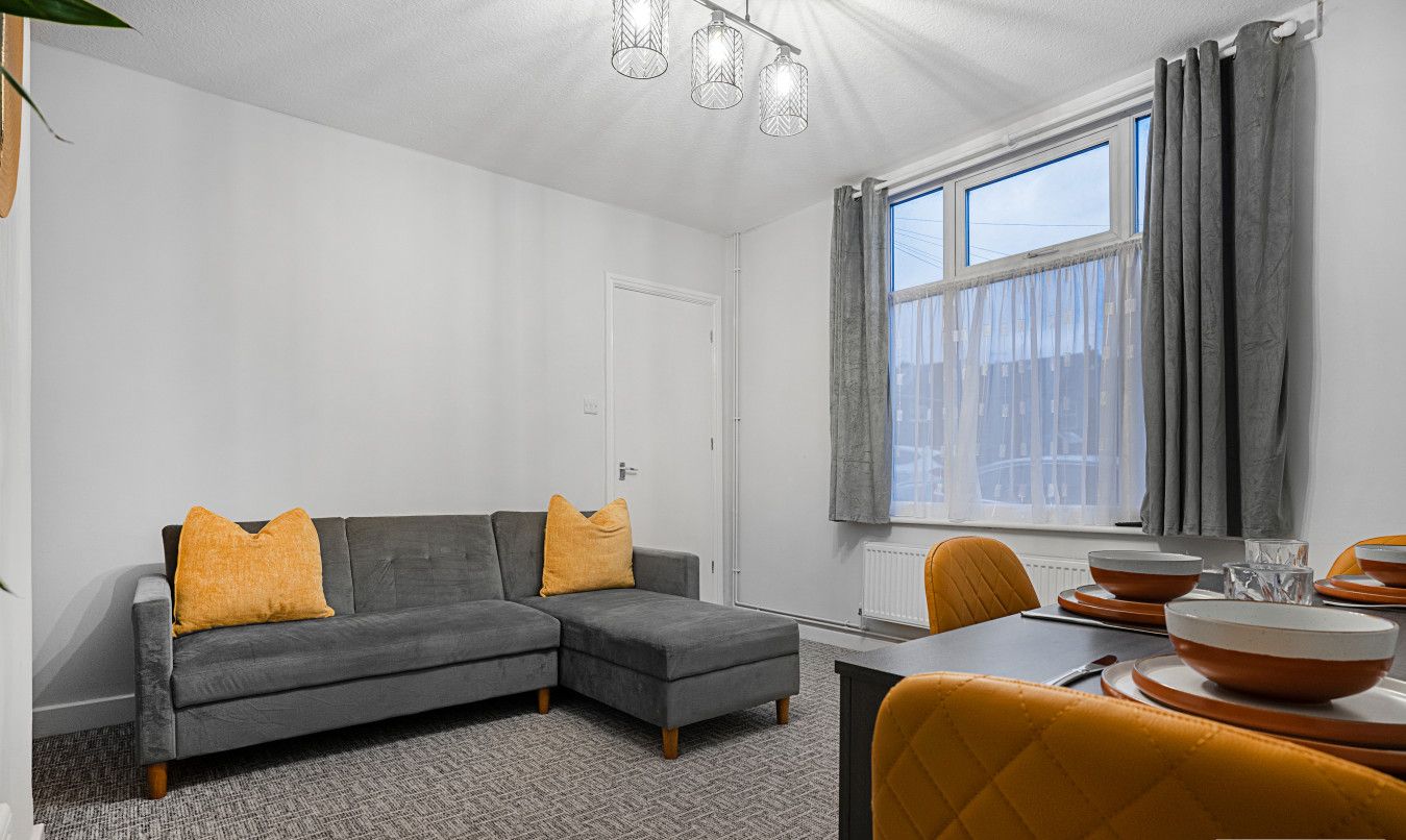 Un salon moderne avec un canapé d’angle gris, deux coussins de couleur moutarde, une table à manger avec des chaises orange et une fenêtre avec des rideaux gris.