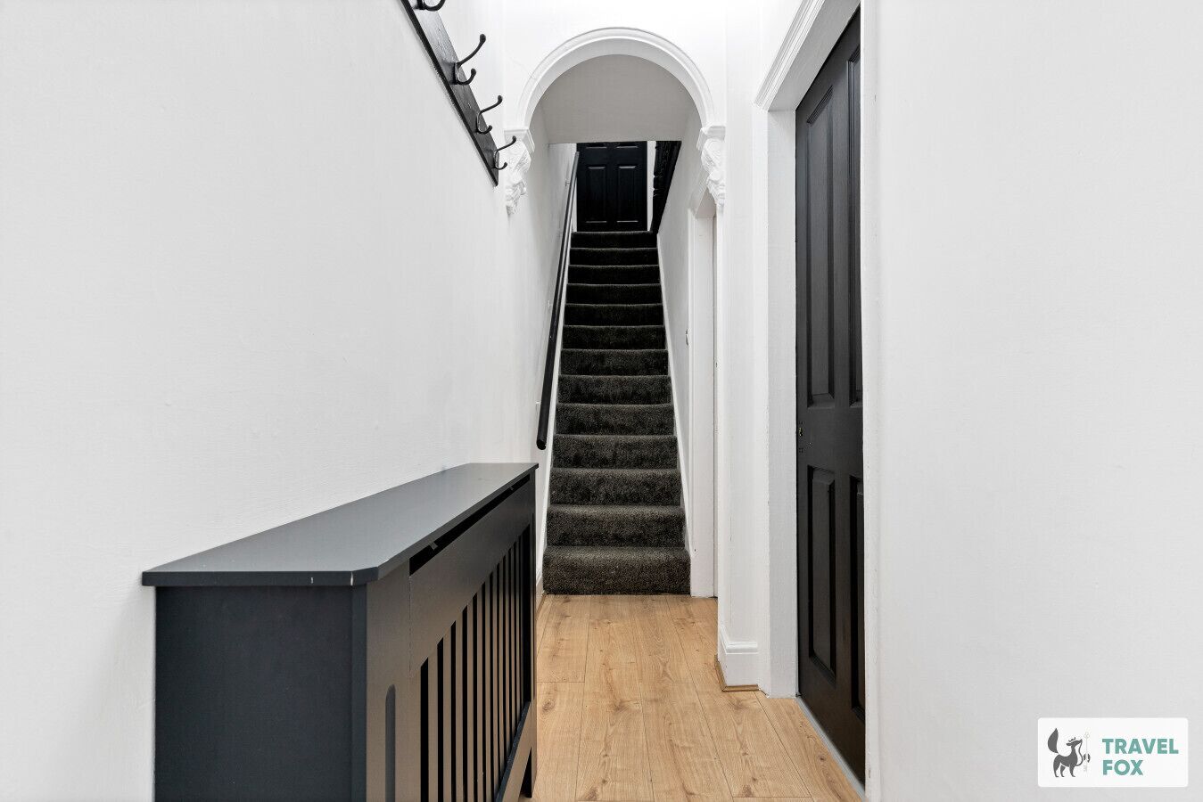 A narrow hallway with a wooden floor, a black railing, and a staircase leading to a dark door.