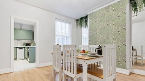 Zona pranzo con tavolo in legno e sedie bianche, cucina con piano di lavoro verde e carta da parati a motivi floreali su una parete.