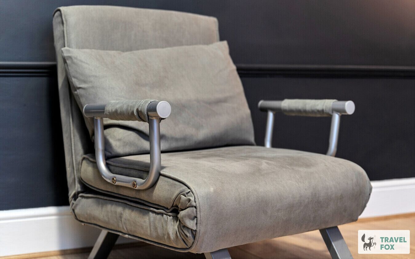 Een grijze bekleden fauteuil met metalen armleuningen en een kussen, geplaatst tegen een donkere muur.
