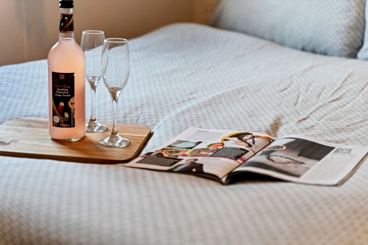 Uma cama com uma revista, uma garrafa de ginger ale gaseificado e dois copos sobre uma bandeja de madeira.