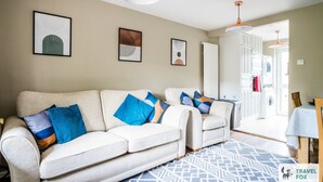 A living room with a beige sofa, blue and orange cushions, a patterned rug, and a washing machine visible through an open door. - Lily's Stay Central  Parking (Nottingham)