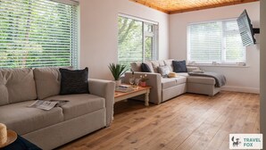 A modern living room with a wooden ceiling, large windows with blinds, a beige sofa, a smaller sofa, and a wooden coffee table. - Orchard Cottage and Hot Tubs (Snowdonia & North Wales)