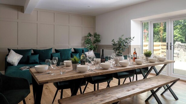 A dining area with a wooden table set for four, surrounded by dark green upholstered chairs. - Orchard Cottage and Hot Tubs (Snowdonia & North Wales)