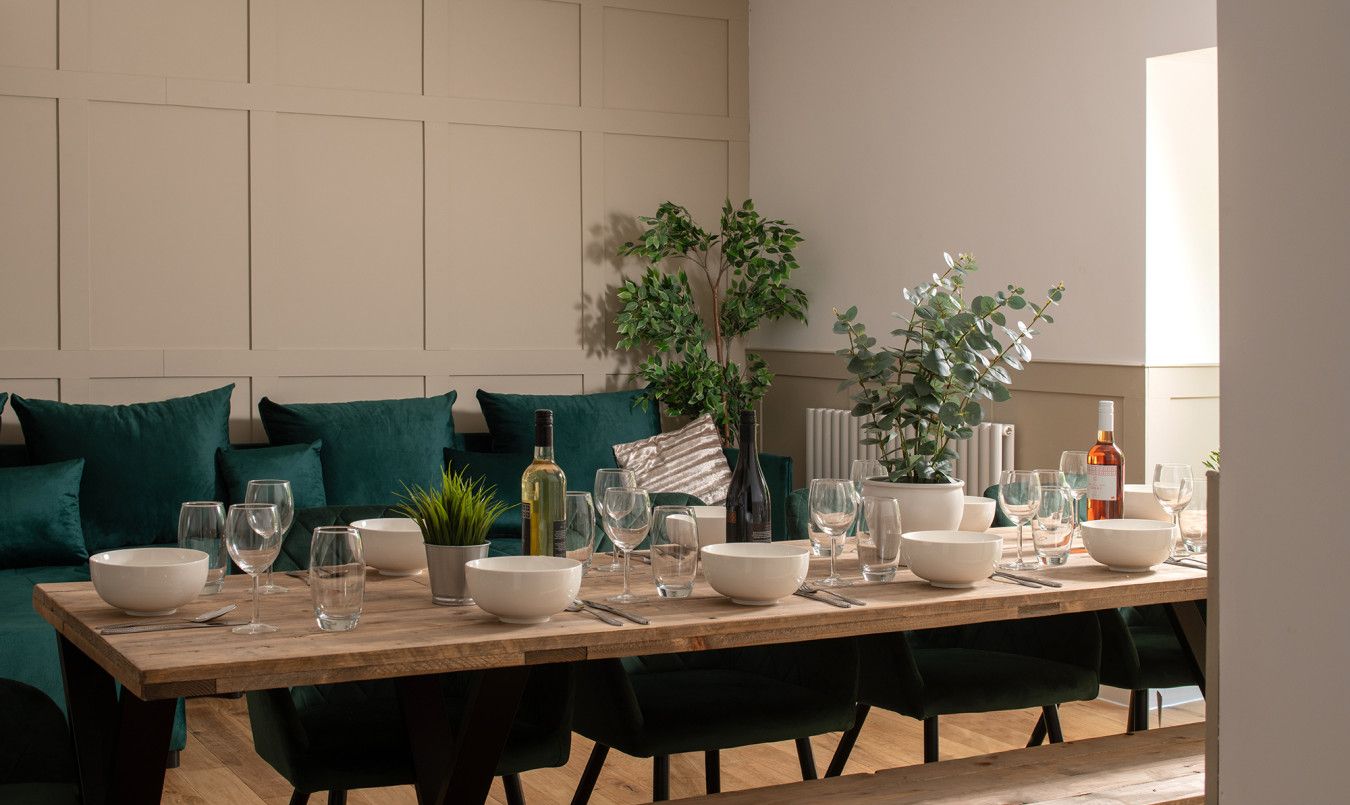 A dining area with a wooden table set for six, surrounded by dark green upholstered chairs. There are several white bowls, wine glasses, and a bottle of wine on the table.