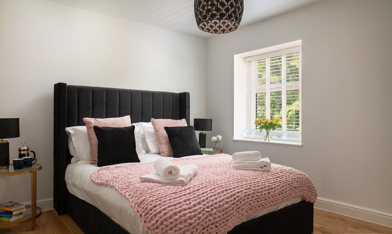 Une chambre à coucher moderne avec une tête de lit noire recouverte de tissu, un plaid rose tricoté et une table de chevet avec une lampe et des livres.
