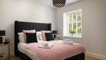 Une chambre à coucher moderne avec une tête de lit noire recouverte de tissu, un plaid rose tricoté et une table de chevet avec une lampe et des livres.