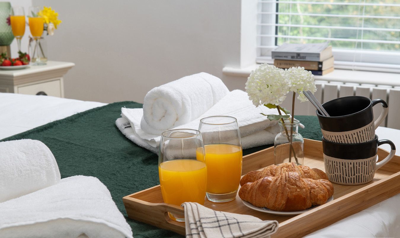 A breakfast tray with croissants, orange juice, and a mug, towels, and a vase with flowers on a bed.
