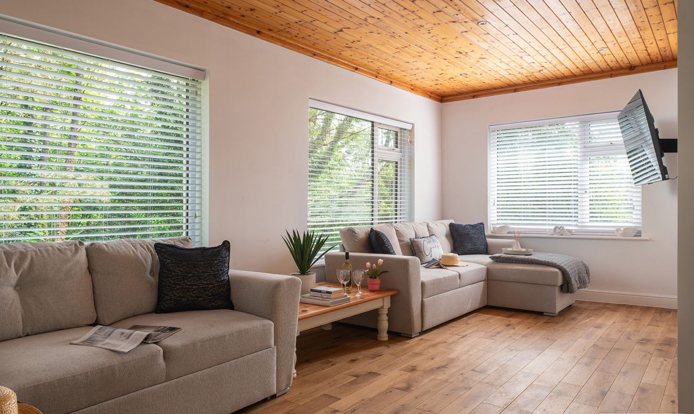 Un soggiorno moderno con soffitto in legno, ampie finestre con persiane, un divano beige, un divano più piccolo e un tavolino in legno.