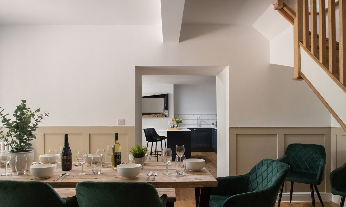 Een eetgedeelte met een houten tafel voor zes personen, omringd door donkergroene gestoffeerde stoelen. Op de tafel staan een potplant en een fles wijn. Achtergrond toont een keuken met een modern design.